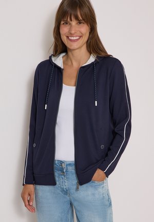 Femme souriante portant un sweat à capuche zippé bleu marine avec un passepoil blanc sur les manches, une doublure de capuche grise, une chemise blanche et un jean bleu clair, une main dans la poche.