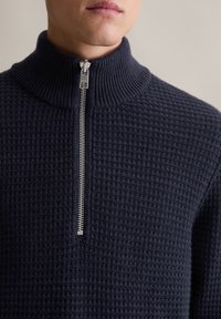 Granatowy sweter dzianinowy z wysokim kołnierzem, z pełnym zamkiem błyskawicznym i teksturowanym wzorem, wykonany z miękkiego, elastycznego materiału.