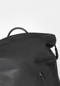 Zaino nero realizzato in tessuto liscio con una parte superiore arrotondata, dotato di una cerniera laterale e di una maniglia in pelle nella parte superiore.