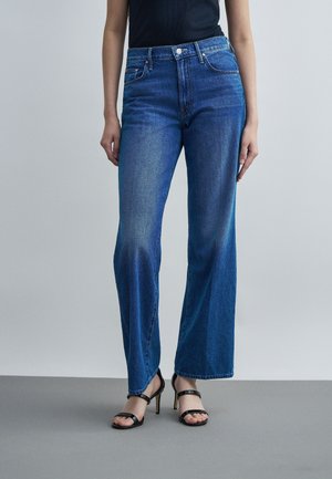 Žena v modrých džínsach so širokými nohavicami a čiernych sandáloch na vysokom opätku stojaca pred jednofarebným svetlošedým pozadím.