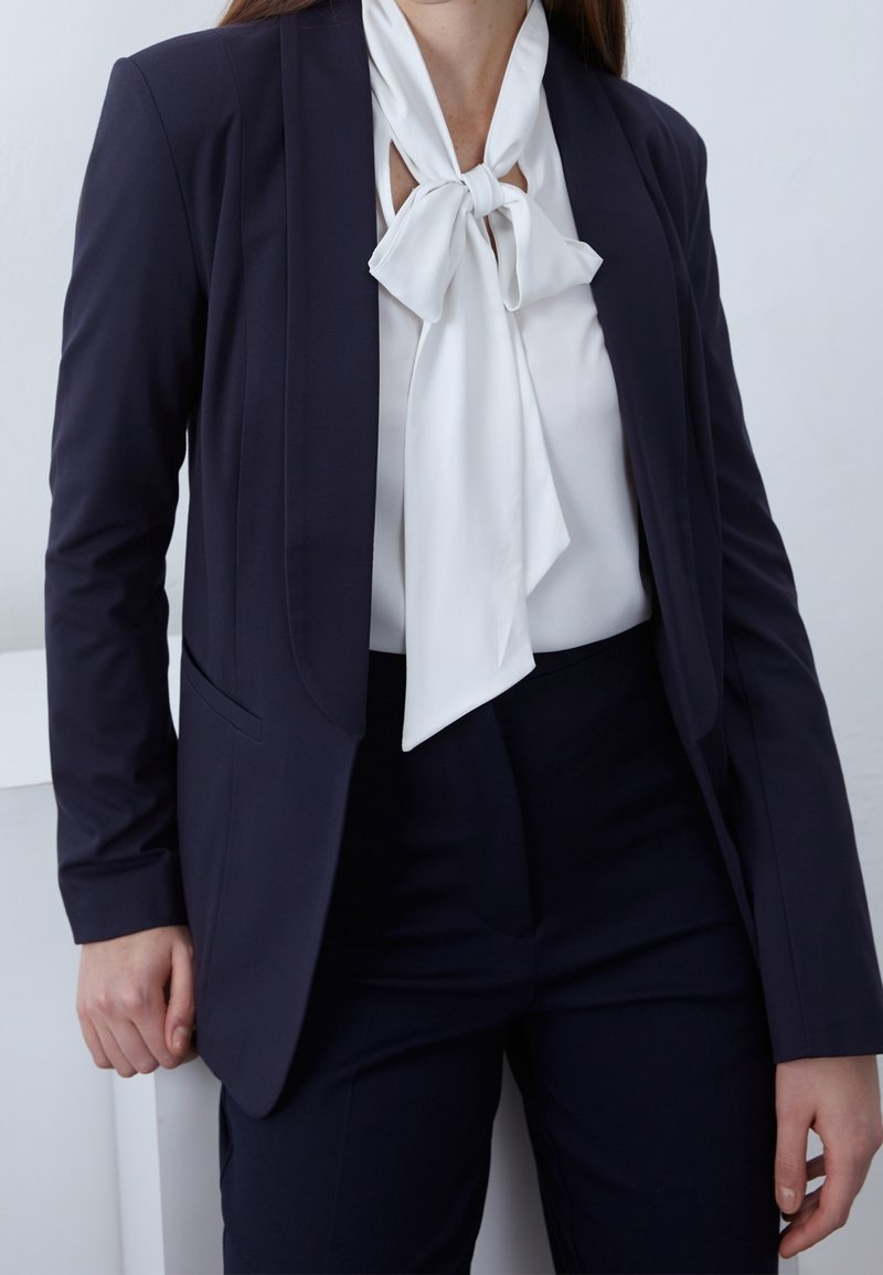 Blazer ajusté bleu marine avec une texture lisse, associé à un chemisier blanc orné d'un nœud papillon au col. Un pantalon ajusté complète la tenue.