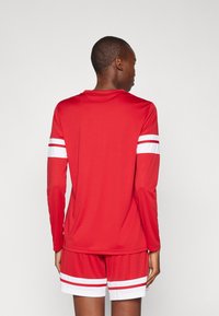Maglietta sportiva rossa a maniche lunghe con strisce bianche sulle maniche, abbinata a pantaloni corti rossi con bande bianche. Tessuto liscio e leggero.