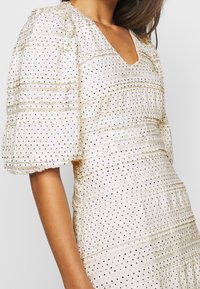 Vestido de manga abullonada en tela crema con patrones texturizados y lunares. Presenta un escote en forma de V y acentos con volantes en capas.