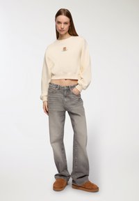 Felpa cropped color crema con design grafico, jeans grigi a vita alta e scarpe marroni slip-on, caratterizzati da una vestibilità rilassata e una texture morbida.