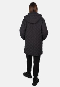 Černá prošívaná péřová bunda s kapucí, dlouhého střihu a strukturovaným vzorem. Nosená s černými kalhotami a teniskami. Pohled zezadu.
