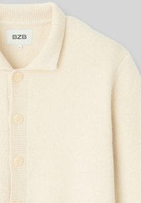Cardigan tricoté de couleur crème avec un col côtelé, comportant cinq boutons ronds et une texture douce. L'étiquette indique "Bzb" et la taille "L."