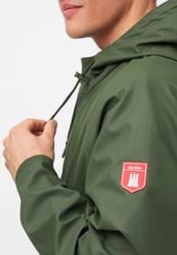 Giacca impermeabile verde con cappuccio, dotata di un patch con logo rosso sul braccio sinistro. Il materiale è liscio con una finitura opaca.