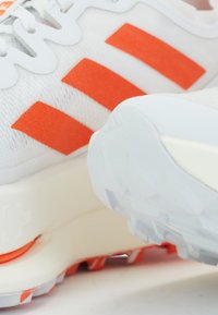 White athletic shoes featuring three orange stripes, textured fabric upper, and a white sole with accent details and a distinct tread pattern.