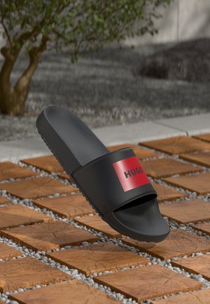 Črne natikače z rdečim logotipom "HUGO" na trakcu, postavljene na lesene ploščice in prodnato površino na prostem, blizu drevesa.