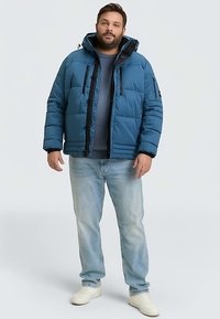 Blauwe donsjas met een zwarte ritssluiting, voorzien van een capuchon en meerdere zakken. Draagt over een grijze trui, gecombineerd met lichte spijkerbroeken en witte schoenen.