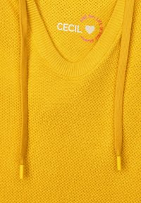 Gelber gestrickter Pullover mit V-Ausschnitt, ausgestattet mit Bindebändern mit kleinen gelben Spitzen und einem weißen Logo am Kragen.