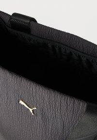 Pochette texturée noire avec un logo Puma doré, partiellement ouverte montrant un intérieur noir avec une poche zippée.