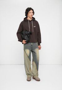 Mlad človek v temno rjavem hoodieju, v jeans hlačah z različnih kosov, s sivimi čevlji, ki drži črno torbo, stoji ob enobarvni beli ozadju.