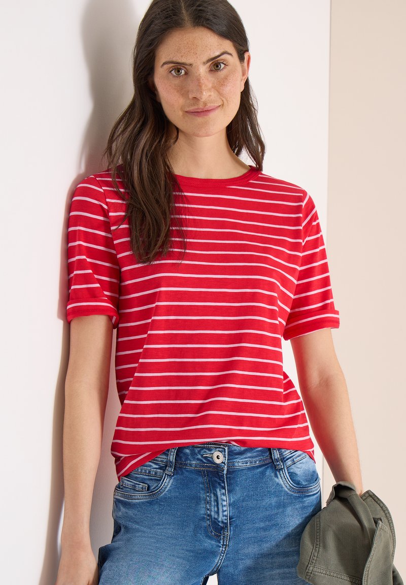 Mujer con cabello largo y oscuro, vestida con camiseta de rayas rojas y blancas y pantalones vaqueros azules, sosteniendo una chaqueta verde, situada frente a un fondo claro.
