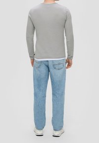 Homme portant un jean bleu clair, des baskets blanches et un t-shirt à manches longues gris, se tenant de dos face à un fond uni.