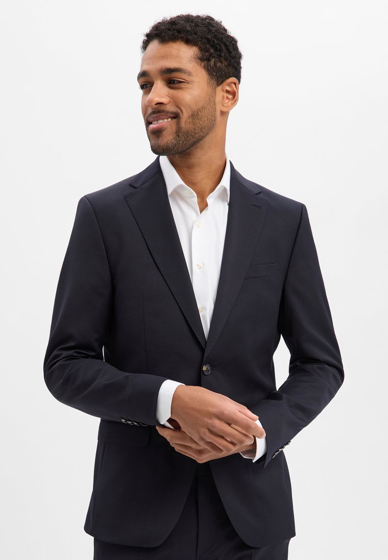 Marineblauer Blazer aus glattem Stoff. Ausgestattet mit einem Reverskragen mit Kerbe, Einzelknopfverschluss und Pattentaschen. Kombiniert mit einem weißen Hemd.