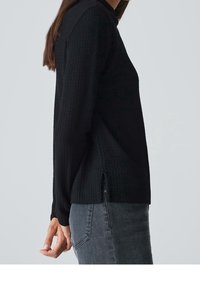 Haut noir texturé à manches longues avec fentes sur les côtés, col rond et coupe décontractée. Associé à un jean gris foncé.