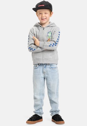 Grauer Zip-Hoodie mit blauen Karomustern und einem gedruckten Design; hellblaue Jeans; schwarze Sneaker mit braunen Sohlen.
