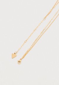 Κολιέ με στρώσεις σε χρυσαφί απόχρωση με τριγωνικό μενταγιόν χαραγμένο με τη λέξη "Guess", σχεδιασμένο με υφασμένες αλυσίδες και κούμπωμα τύπου αστακού.