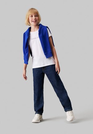 Camiseta blanca con el texto "Calvin Klein", sudadera azul caída sobre los hombros, jeans azul oscuro y zapatillas blancas. Niño pequeño con el cabello corto.
