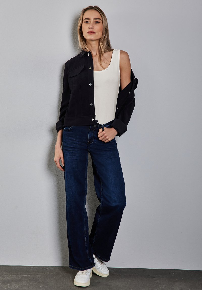 Chaqueta negra corta sobre una camiseta blanca de tirantes, combinada con jeans de pierna ancha azules y zapatillas blancas. Textura suave, diseño casual.