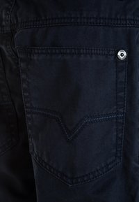 Námořnická denimová kapsa s dekorativním V-šitím, stříbrným knoflíkem na zapínání a kontrastním modrým prošíváním.