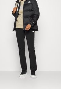 Doudoune noire sur un sweat à capuche beige, assortie à un pantalon noir avec des logos et des baskets noir et blanc. Design moderne pour l'extérieur.