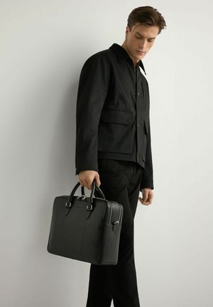 Mężczyzna w czarnej kurtce i spodniach trzymający czarną skórzaną teczkę, stojący na tle jasnego, jednolitego tła.