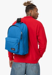 Niebieski plecak z materiału, z przednią kieszenią na zamek, zaokrąglonym dnem i czarnymi regulowanymi paskami, noszony na czerwonej bluzie.
