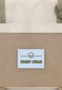Tessuto color beige con una superficie testurizzata, caratterizzato da un'etichetta bianca con "JOHNNY URBAN" in testo verde e un anello di accento olive nella parte superiore.