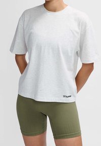 Camisa blanca de manga corta con textura moteada, cuello redondo y logo de Hummel en el dobladillo, combinada con pantalones cortos de ciclismo verdes.