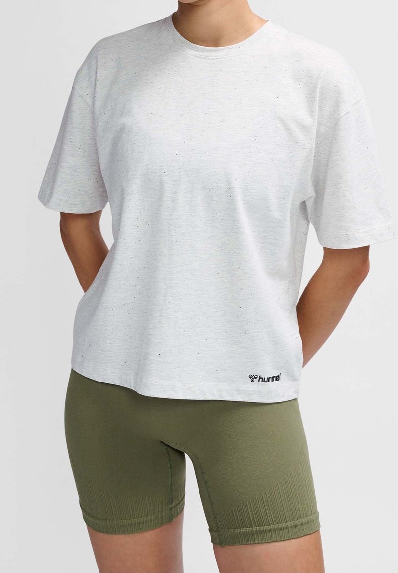 Camisa blanca de manga corta con textura moteada, cuello redondo y logo de Hummel en el dobladillo, combinada con pantalones cortos de ciclismo verdes.