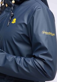 Marinblå, vattentät jacka gjord av ett slätt material; har en gul logotyp på bröstet och trädetaljer på dragsnörena.