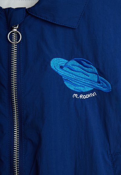 Μπλε μπουφάν με ασημί φερμουάρ και κεντημένο μπλε πλανήτη με δαχτυλίδια και το κείμενο «M. RODINI» κάτω από την περιοχή του στήθους.
