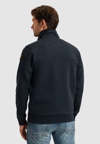 Marineblaue Sweatshirtjacke mit hohem Kragen, gerippten Bündchen und Saum sowie Nahtdetails auf der Rückseite. Mit einem Logo-Patch an der linken Schulter.