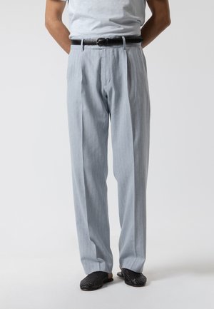 Hombre con pantalones plisados gris claro y zapatillas negras tejidas sin cordones, con cinturón negro, de pie con las manos detrás de la espalda.