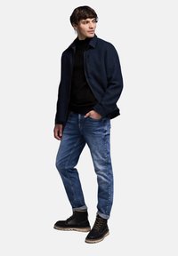 Mörkblå jacka med en dragkedjeficka, svart texturerad tröja, ljusblå jeans och svarta stövlar med beige sulor och snören.