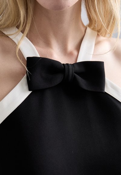 Svart halterneck med hvit kant og en sløyfedetalj ved halsen. Stoffet ser glatt ut med en matt finish.