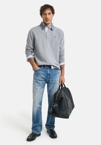 Grauem Pullover mit Kragen und blauen Akzenten, darunter ein hellblaues Hemd, blaue Jeans, schwarze Schuhe und eine schwarze Sporttasche.