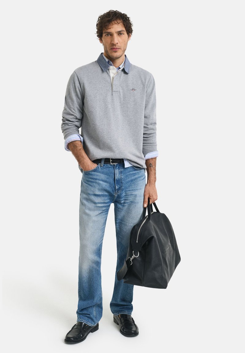 Suéter gris con cuello y acentos azules, camisa celeste debajo, vaqueros azules, zapatos negros y una bolsa de viaje negra.
