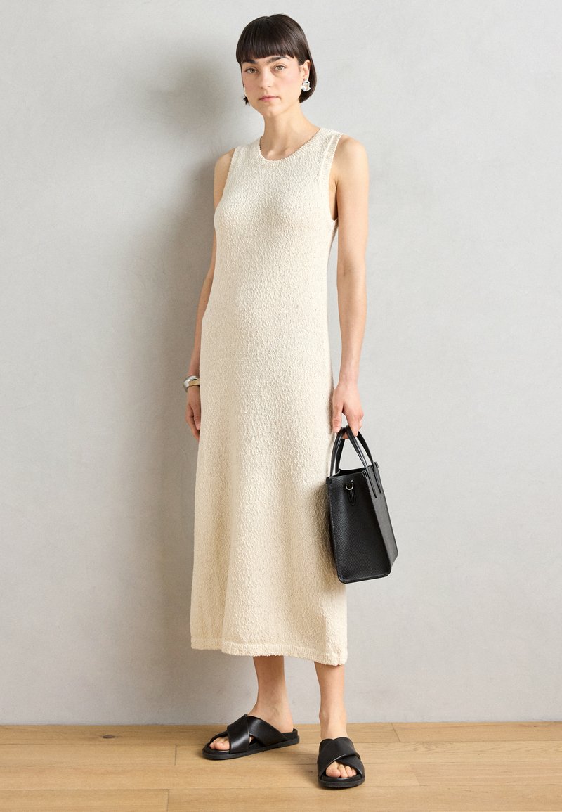 Robe crème sans manches, longueur genoux avec un tissu texturé. Associée à des sandales noires à enfiler et un petit sac à main noir. Design lisse et minimal.