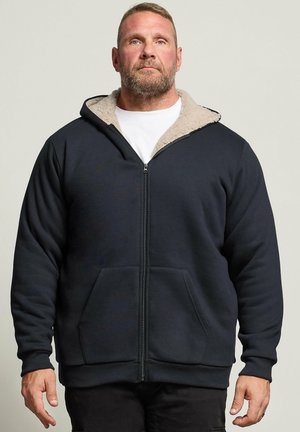 Homme barbu portant une veste polaire noire zippée avec une chemise blanche en dessous, debout devant un fond clair uni.