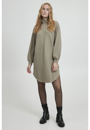 Khaki lange mouwen jurk met een halve ritssluiting kraag, textuurstof en een rond afgewerkte zoom; gecombineerd met zwarte enkellaarsjes en doorschijnende panty's.