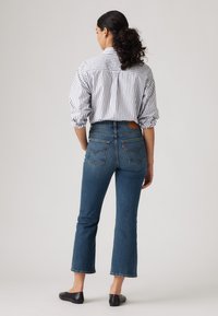 Donna con capelli ricci raccolti in una coda di cavallo, vestita con una camicia a righe bianche e nere infilata in jeans blu a tre quarti e ballerine nere, in piedi di spalle.