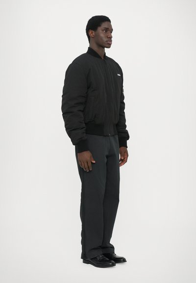 Schwarze Bomberjacke mit gerippten Bündchen und Kragen, kombiniert mit dunklen Hosen und schwarzen Schuhen. Verfügt über einen Reißverschluss und Seitentaschen.