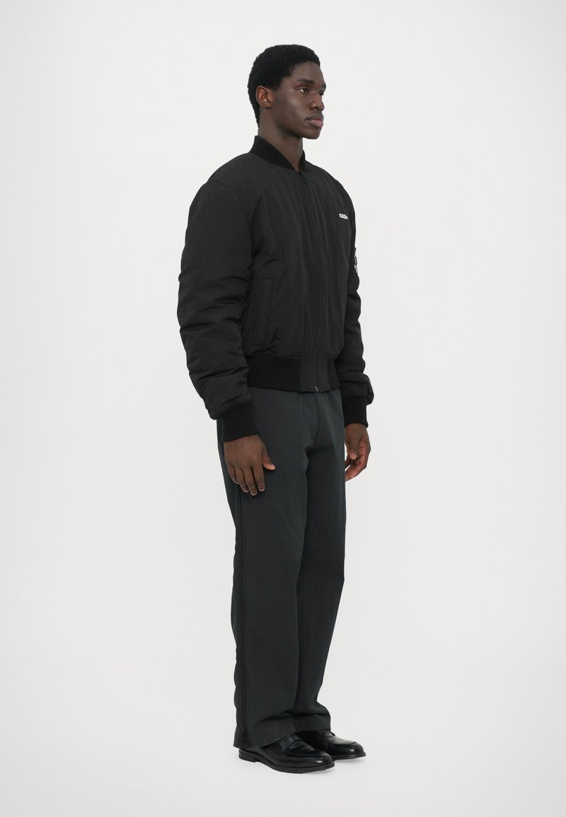 Chaqueta bomber negra con puños y cuello acanalados, combinada con pantalones oscuros y zapatos negros. Presenta un cierre de cremallera y bolsillos laterales.