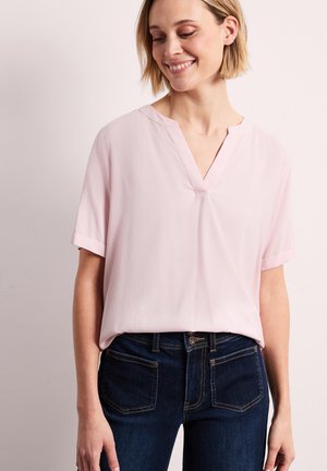 Femme portant un chemisier rose pâle à manches courtes et col en V, associé à un jean en denim bleu foncé, souriant doucement en regardant vers le bas.