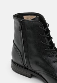 Botín de cuero negro hasta el tobillo con un acabado texturizado, diseño de cordones, cremallera lateral y un tacón apilado para mayor estabilidad.