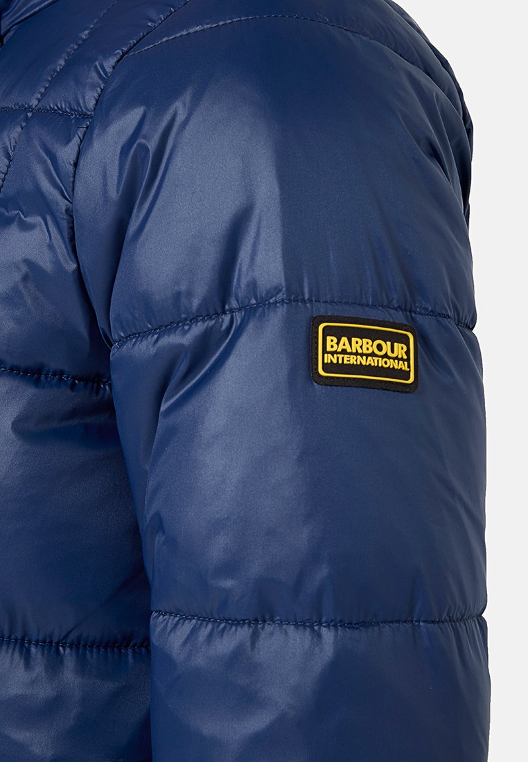 Giacca puffer blu navy con design trapuntato, caratterizzata da una toppa in gomma gialla con il logo "Barbour International" sulla manica. Texture morbida e lucida.