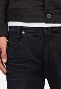 Černé džíny z denimu s hladkou texturou, vybavené pětinásobným designem kapes a kovovými detaily. Nosené s bílou košilí pod nimi.